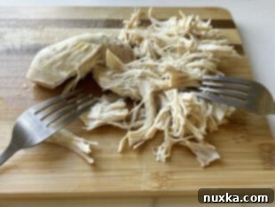 Cooked chicken breasts being shredded with two forks inside a bowl, ready to be added back to the white chicken chili.