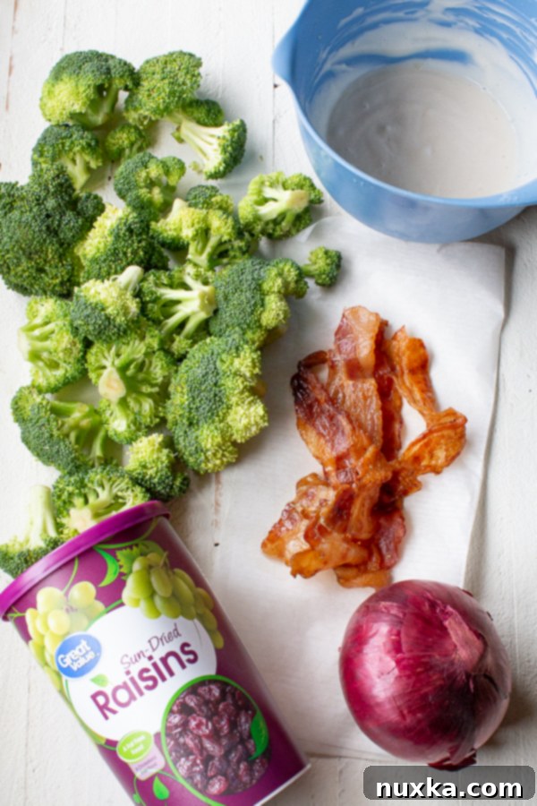 A flat lay of fresh ingredients for broccoli salad including a head of broccoli, bacon, red onion, and raisins.