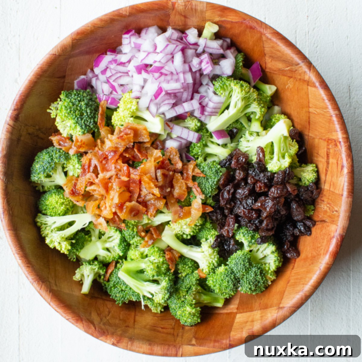 Prepped ingredients for broccoli salad including chopped broccoli, bacon, onions, and raisins in separate sections of a bowl.