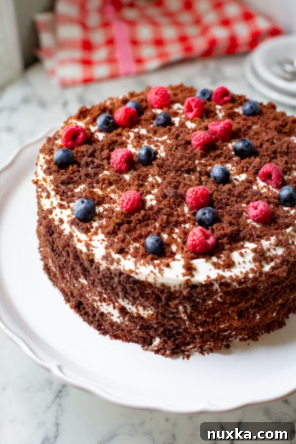 A two-layer chocolate cake with a thick layer of white cream cheese frosting, topped with fresh blueberries, raspberries, and a sprinkle of chocolate cake crumbs. The cake is perfectly moist and inviting.