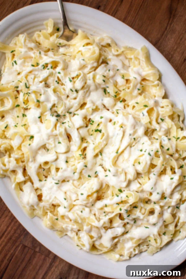 super creamy and rich fettuccine Alfredo sauce smothered in fettuccine pasta and parsley flakes on a white serving dish with a fork