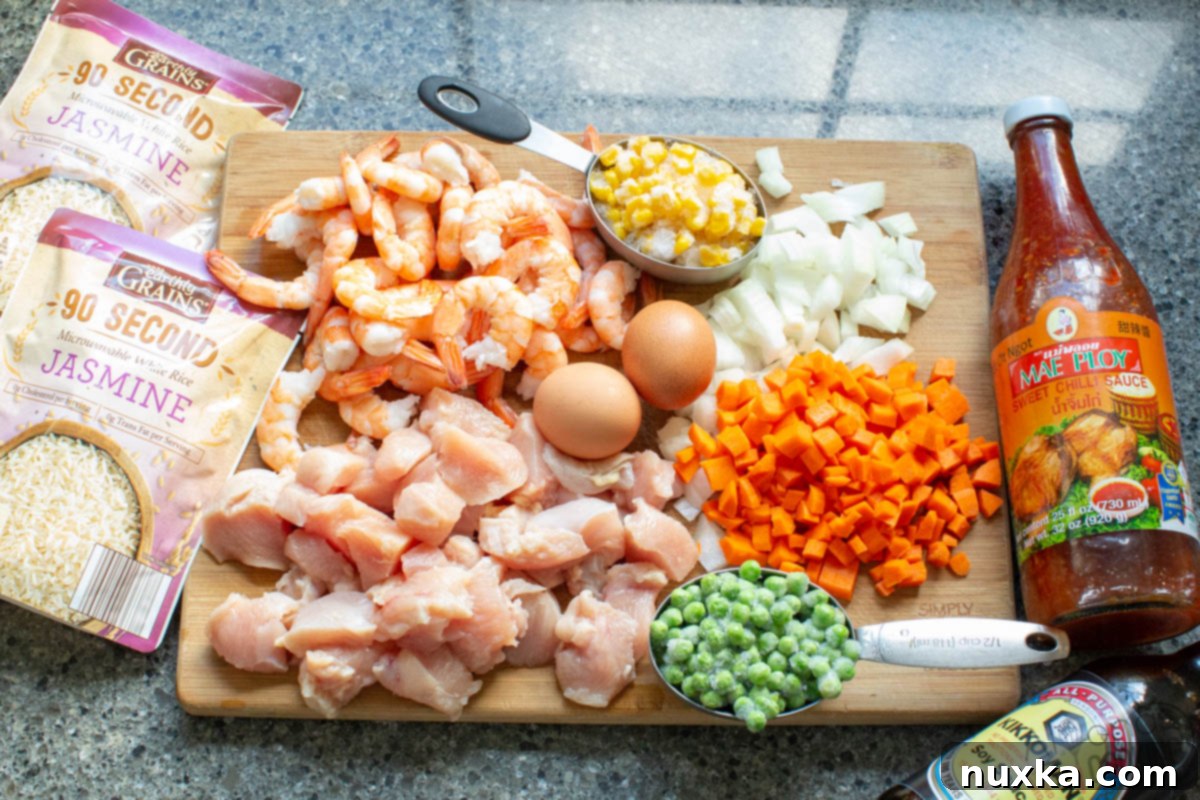 All the fresh ingredients for homemade Shrimp Fried Rice, including cooked rice, chicken breasts, cooked shrimp, frozen peas, corn, diced carrots, onion, eggs, Kikkoman soy sauce, Mae Ploy sweet chili sauce, and cooking oil.