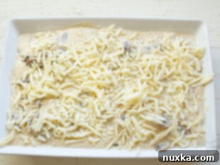 Mushroom Alfredo sauce being poured over sliced potatoes in a casserole dish