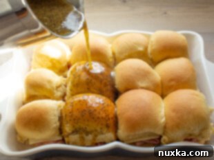 Hot poppy seed sauce being poured over assembled ham and cheese sliders.