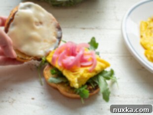 Assembling an egg sandwich with layers of arugula, scrambled egg patty, pickled red onion, and grainy mustard on a brioche bun