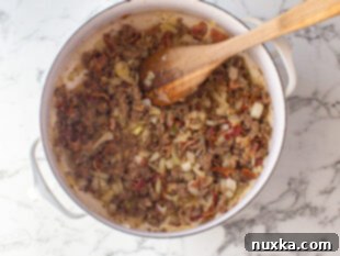 Close-up of a large pot on a stovetop, showing diced onions, crumbled Italian sausage, and chopped bacon sautéing, forming the base for Zuppa Toscana.