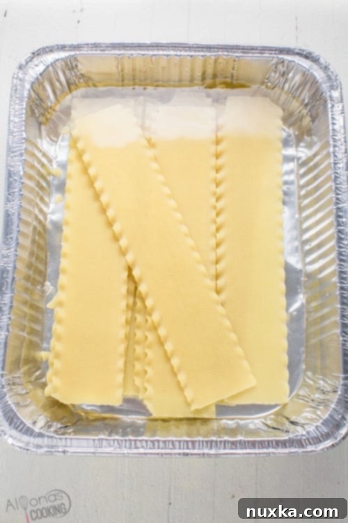 Soaked lasagna noodle sheets arranged in a disposable aluminum pan filled with hot water, ready for layering.